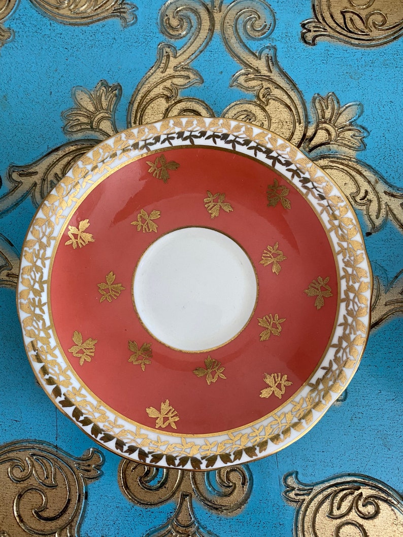May include: A coral-colored saucer with a gold rim and a delicate floral pattern. The saucer is made of fine china and has a white center.