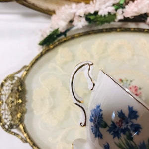 May include: A white teacup with a gold rim and a blue floral pattern. The teacup is sitting on a white and gold tray.