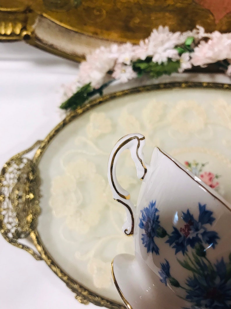 May include: A white teacup with a gold rim and a blue floral pattern. The teacup is sitting on a white and gold tray.