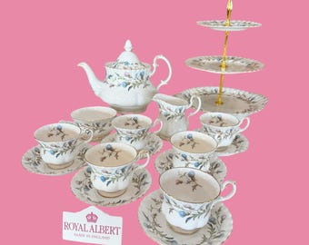 Könnte beinhalten: Ein Royal Albert Teeservice bestehend aus einer weißen Teekanne, einem Milchkännchen und sechs Tassen mit passenden Untertassen, verziert mit blauen und grünen Blumenmustern. Ein dreistöckiger Servierständer mit goldfarbenem Griff ist enthalten.