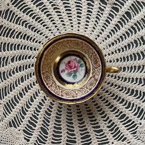 May include: A vintage teacup with a blue and gold rim and a floral design in the center. The cup is sitting on a white crocheted doily.