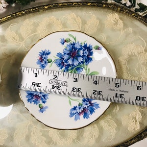 May include: A white saucer with a gold rim and a floral design featuring blue and purple cornflowers. The saucer is 5 inches in diameter.