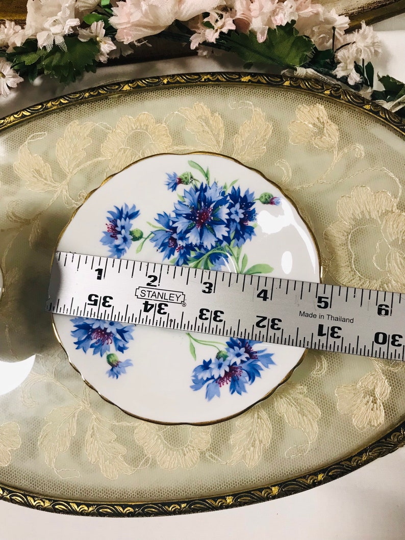 May include: A white saucer with a gold rim and a floral design featuring blue and purple cornflowers. The saucer is 5 inches in diameter.