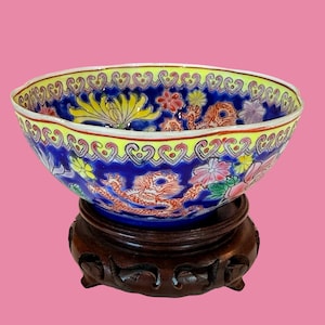 May include: A decorative, oval-shaped ceramic bowl with a blue interior, yellow rim, and floral and dragon motifs. The bowl sits on a dark brown wooden stand. The bowl's exterior features a detailed design with red, pink, and yellow flowers.