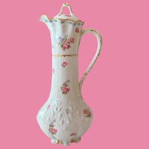 May include: A tall white porcelain chocolate pot with a lid, featuring a delicate floral pattern of pink roses and blue flowers. The vessel has a fluted body, a curved handle, and gold trim accents along the rim, base, handle, and lid finial.