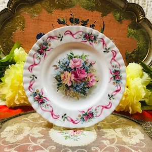 Puede incluir: Un plato de porcelana blanca con un diseño floral rosa y azul. El plato presenta un ramo de rosas, nomeolvides y otras flores con cintas rosas alrededor del borde.