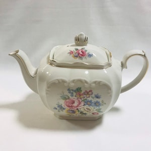 May include: A white teapot with a floral design and gold trim. The teapot has a square body and a lid with a knob on top. The floral design features pink, yellow, and blue flowers.