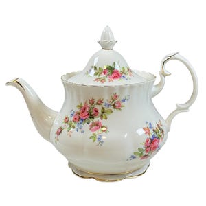 Puede incluir: Una tetera de porcelana blanca con un diseño floral de rosas rosas y flores azules. La tetera tiene un pico curvo, un asa y una tapa con un adorno decorativo. Ribete dorado.