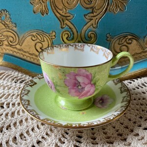 May include: A green teacup and saucer with a floral design. The cup and saucer have a gold trim and are decorated with pink and white flowers.