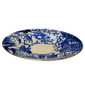 Könnte beinhalten: Ovale Servierplatte aus Keramik mit blau-weißem Landschaftsdesign. Der Rand ist mit einem goldenen Band verziert. Die Platte zeigt eine detaillierte Illustration von Bäumen, Gebäuden und Figuren.