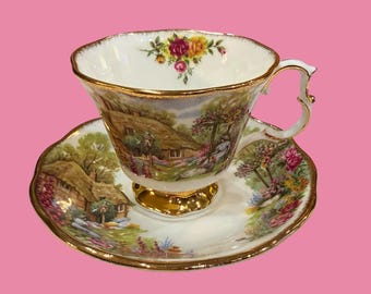 May include: A white teacup and saucer set with a gold rim and foot. The cup and saucer feature a pastoral scene with a cottage and flowers. A floral arrangement decorates the top of the cup. The set is on a pink background.
