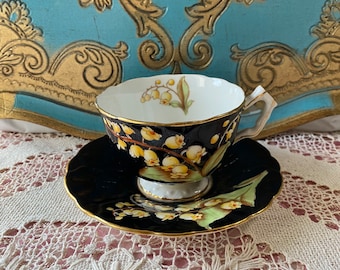 Könnte beinhalten: Ein schwarzes und weißes Tee-Set mit einem floralen Design aus gelben Lilien. Tasse und Untertasse haben einen goldenen Rand.