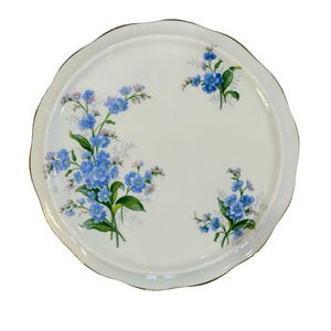 May include: A white, scalloped-edge serving plate with a gold rim. The plate is decorated with clusters of blue and pink forget-me-not flowers and green leaves. The floral design is spread across the plate's surface.