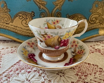 Op de afbeelding: Een vintage theekopje en schotelset met een bloemenpatroon met peren, druiven en bloemen in tinten geel, rood en bruin. De set heeft een gouden rand en is gemaakt van fijn bottenporselein.