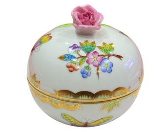 May include: A white porcelain trinket box with a pink rose on the lid. The box is decorated with colorful floral and butterfly designs, and gold trim. The box is round and has a lid.