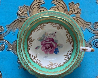 May include: A green and gold teacup and saucer with a floral design. The cup and saucer have a scalloped edge and a gold trim.