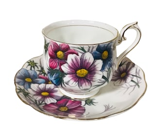 May include: A white teacup and saucer set with a floral design featuring pink, purple, and blue flowers. The cup and saucer have a gold trim.