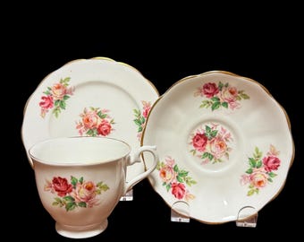 May include: A vintage teacup, saucer, and plate set. The white porcelain pieces feature a floral rose pattern in shades of pink and yellow, with green leaves. The edges are trimmed with gold. The set includes a teacup, a saucer, and a plate.