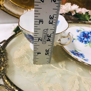 May include: A white teacup and saucer with a floral pattern featuring blue flowers. The teacup and saucer are sitting on a gold-colored tray with a white lace overlay. A ruler is visible in the background, indicating the teacup is approximately 3.5 inches tall.