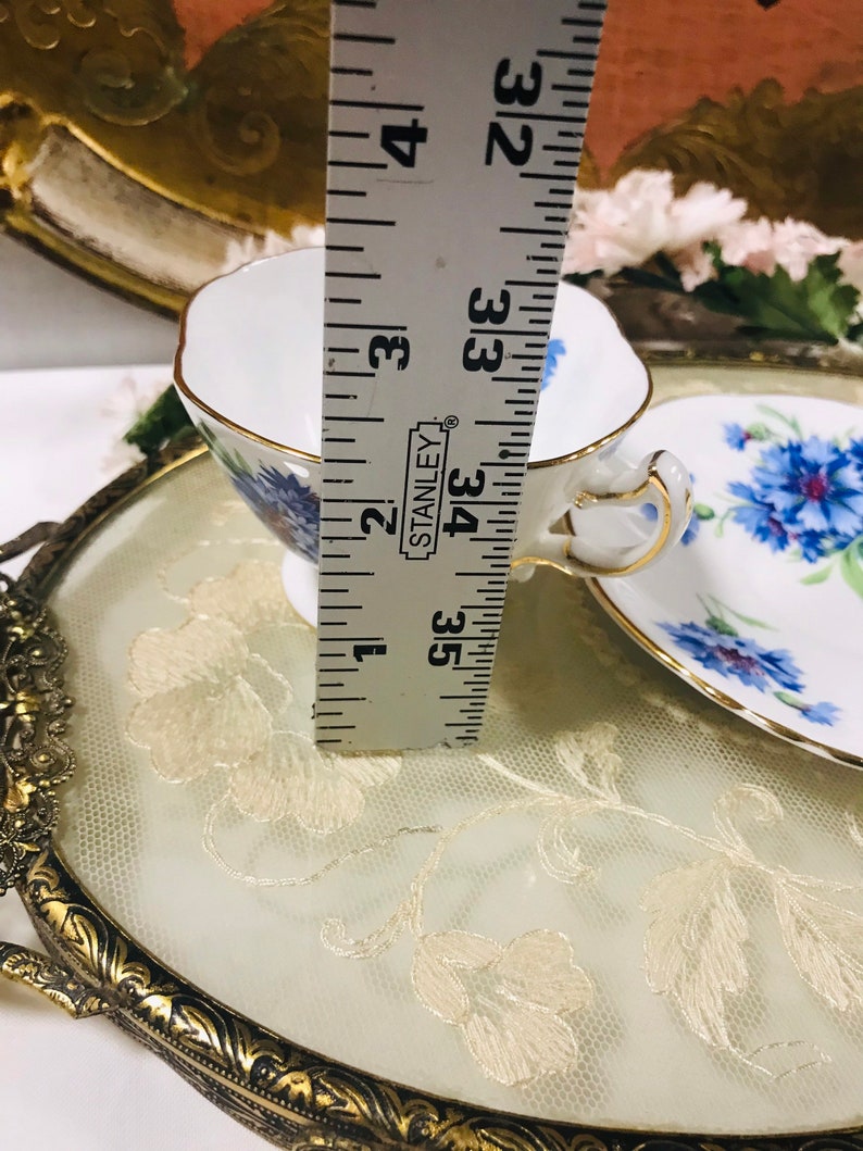 May include: A white teacup and saucer with a floral pattern featuring blue flowers. The teacup and saucer are sitting on a gold-colored tray with a white lace overlay. A ruler is visible in the background, indicating the teacup is approximately 3.5 inches tall.
