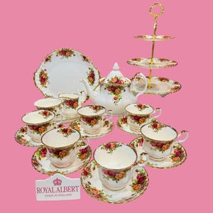 May include: A Royal Albert tea set featuring white porcelain with red and yellow rose patterns and gold trim. The set includes a teapot, a three-tier cake stand, a creamer, and multiple teacups with matching saucers. A sign reads ROYAL ALBERT MADE IN ENGLAND.
