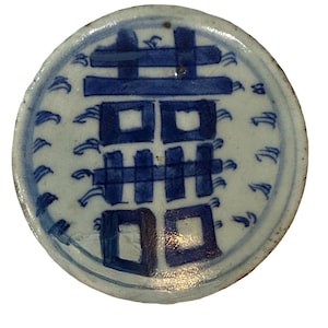 May include: A circular ceramic plate featuring a white base with a hand-painted blue double happiness character in the center. The design is surrounded by small, stylized blue wave patterns along the outer edge of the plate's surface.
