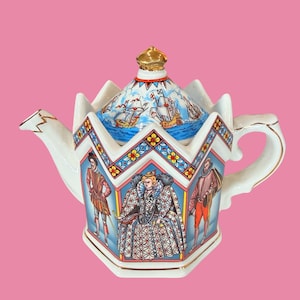 May include: A hexagonal ceramic teapot featuring a crown-shaped lid with a gold finial. The body displays colourful illustrations of historical figures in period attire against a blue sky. The lid depicts sailing ships on a blue sea.