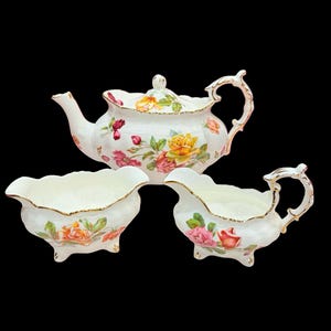 Puede incluir: Un juego de té de porcelana blanca con tetera, jarra de crema y azucarero. El juego está decorado con rosas coloridas y ribetes dorados. La tetera tiene tapa y asa curvada. La jarra de crema y el azucarero tienen borde festoneado y patas pequeñas.