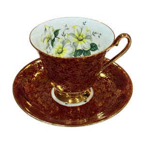 May include: A vintage teacup and saucer set. The teacup and saucer are a deep red with a gold floral pattern. The inside of the teacup is white with a floral design of white and yellow flowers. The handle and base of the teacup are gold.