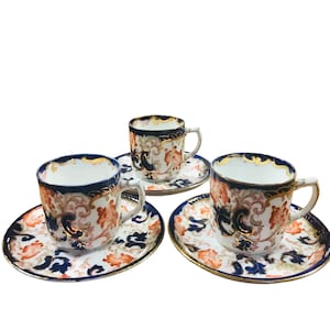 Puede incluir: Juego de tres tazas de té y platillos vintage con un patrón floral en tonos azules, naranjas y dorados. Las tazas y los platillos tienen un fondo blanco con un borde azul.