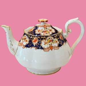 以下が含まれることがあります： 白い磁器製のティーポットで、波打つような形状の本体と花の形をしたつまみ付きの蓋が特徴です。上部には深い青色の帯、精巧な金の装飾、そしてオレンジ、黄色、緑の色合いで手描きされた花の模様が施されています。