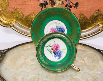 May include: A green teacup and saucer with gold trim and floral designs. The teacup and saucer are on a white oval tray with gold trim and a floral design.