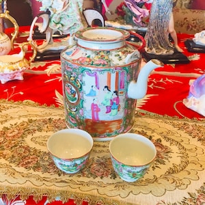 Puede incluir: Una tetera de porcelana con un diseño floral y una escena de personas con ropa tradicional. La tetera está acompañada de dos tazas de té a juego. La tetera y las tazas de té están sobre un mantel de mesa con estampado dorado y crema.