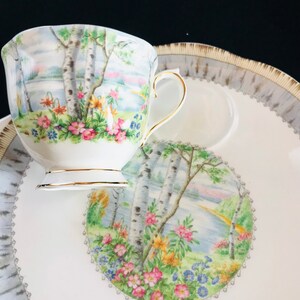 Pu&ograve; includere: Servizio da t&egrave; bianco con un design paesaggistico con betulle, fiori e uno specchio d'acqua. La tazza e il piattino hanno finiture dorate. Il piattino ha un bordo decorativo.