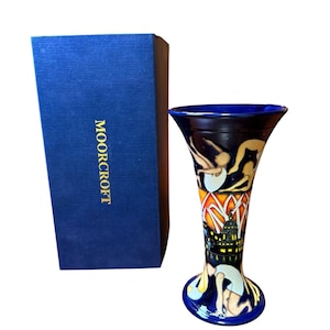 May include: A decorative vase with a flared top and a detailed illustration of figures and a cityscape. The vase is predominantly blue with accents of yellow, orange, and white. A blue box with the word "MOORCROFT" is next to the vase.