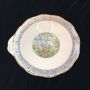 Pu&ograve; includere: Un piatto da portata in ceramica bianca con bordo azzurro e oro. Il centro presenta una scena paesaggistica circolare con alberi, fiori e uno specchio d'acqua. Il piatto ha un manico e un bordo smerlato.