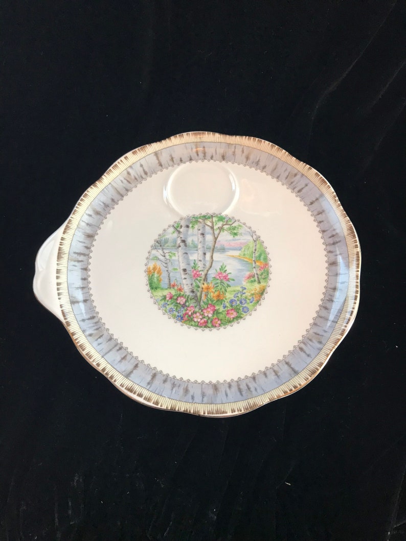 Pu&ograve; includere: Un piatto da portata in ceramica bianca con bordo azzurro e oro. Il centro presenta una scena paesaggistica circolare con alberi, fiori e uno specchio d'acqua. Il piatto ha un manico e un bordo smerlato.