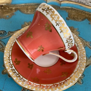 May include: A coral-colored teacup and saucer set with a gold trim and floral pattern. The set is on a blue and gold tray.