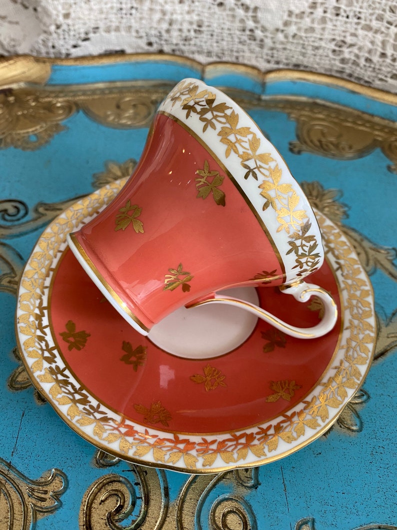 May include: A coral-colored teacup and saucer set with a gold trim and floral pattern. The set is on a blue and gold tray.