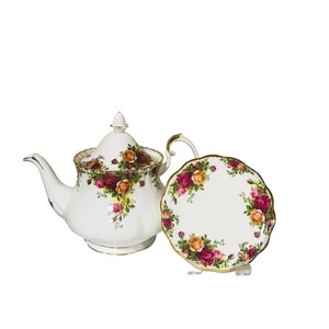 May include: A white teapot and matching saucer with a floral pattern of red, yellow, and pink roses. The teapot has a gold trim and a lid with a knob. The saucer has a scalloped edge.