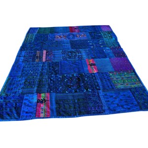Op de afbeelding: Een blauwe patchworkquilt met verschillende patronen en texturen. De quilt is rechthoekig en lijkt handgemaakt, met verschillende stoffen aan elkaar genaaid. Het kleurenpalet omvat diepblauw, indigo en accenten van paars en roze.