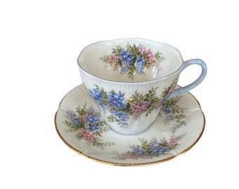 Könnte beinhalten: Eine weiße Teetasse und Untertasse mit einem Blumenmuster in Blau- und Rosatönen. Die Tasse hat einen zarten, gewellten Rand und einen Henkel. Die Untertasse hat ein passendes Blumenmuster und einen goldenen Rand.