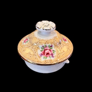 May include: A white ceramic teapot lid with a gold trim and floral design. The lid features a raised floral knob and a delicate pattern of roses and leaves.