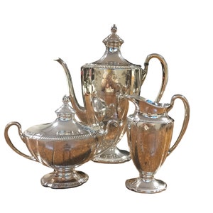 Może przedstawiać: Srebrny zestaw do herbaty z czajnikiem, dzbankiem na śmietanę i cukiernicą. Zestaw jest ozdobiony wzorem kwiatowym.