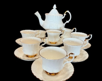 May include: A white porcelain tea set with gold detailing. The set comprises a teapot, creamer, sugar bowl, and six teacups with saucers. The teacups and saucers feature a scalloped edge. The set is presented against a black backdrop.