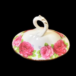 May include: A white ceramic lid with a curved handle and gold trim. The lid is adorned with pink roses and green leaves. The handle features gold detailing. The lid is likely for a serving dish or container.