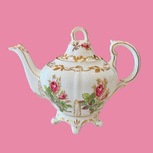Peut inclure: Une théière en porcelaine blanche avec un corps cannelé, un couvercle assorti muni d'une anse en boucle et un bec verseur incurvé. La théière est ornée de motifs de roses roses, de feuilles vertes et de dorures sur le bord, l'anse et la base.
