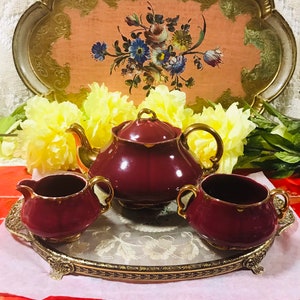 Puede incluir: Un juego de té de color marrón rojizo con una tetera, jarra de crema y azucarero sobre una bandeja dorada con un diseño floral. La bandeja tiene un borde elevado y patas ornamentadas.