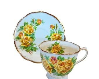 May include: A vintage teacup and saucer set. The cup and saucer are white with a light blue background and adorned with yellow roses and green leaves. The edges are trimmed with gold.