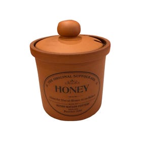 May include: A terracotta-colored honey jar with a matching lid. The jar has a round shape with a slightly flared rim and a small knob on the lid. The jar is labeled "THE ORIGINAL SUFFOLK JAR" and "HONEY".